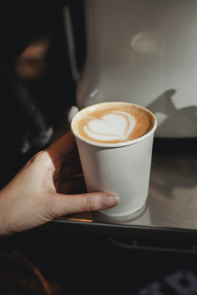 Crop anonymous female touching of aromatic hot cappuccino served in takeaway paper cup in blurred cafe