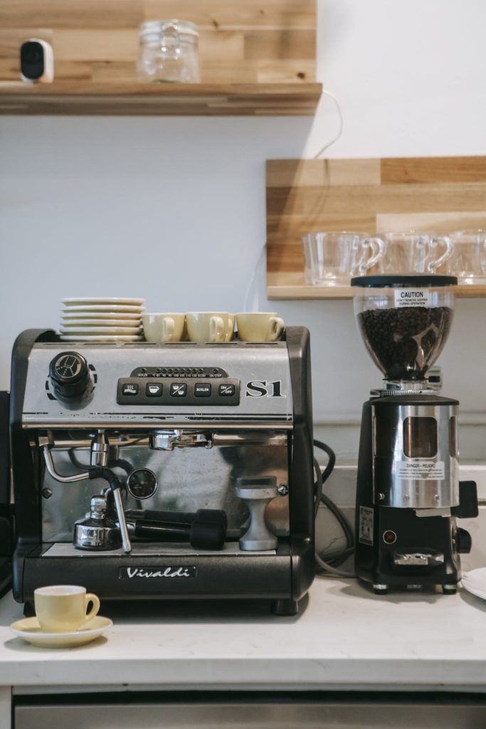 Modern stainless coffee machine placed on counter in kitchen under shelves with coffeemaker and ceramic mugs